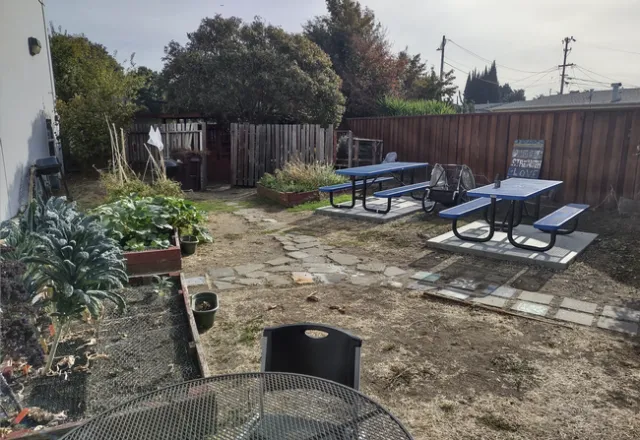 Two blue picnic tables with attached benches sit o concrete slabs among the garden beds. A sign leaning against the fence reads,