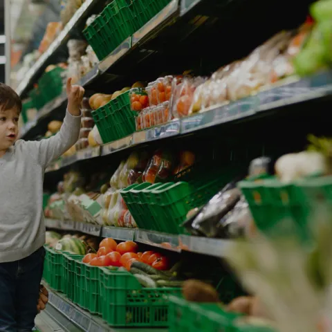 A woman with her young child standing in the produce aisle picking out vegetables. The Nutrition Policy Institute logo is on the right.